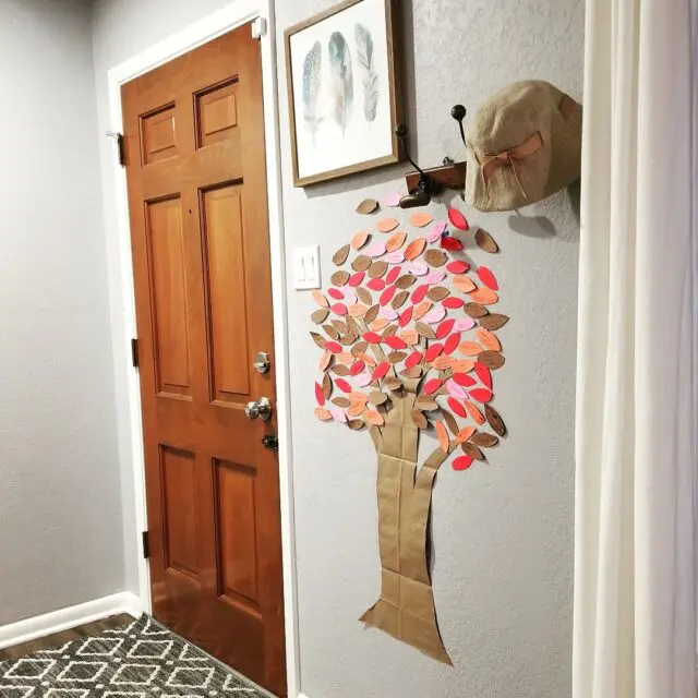 Sideways shot of a paper tree with fall colored leaves taped next to a front door, below a watercolor paining of blue and green feathers and below a hat rack.