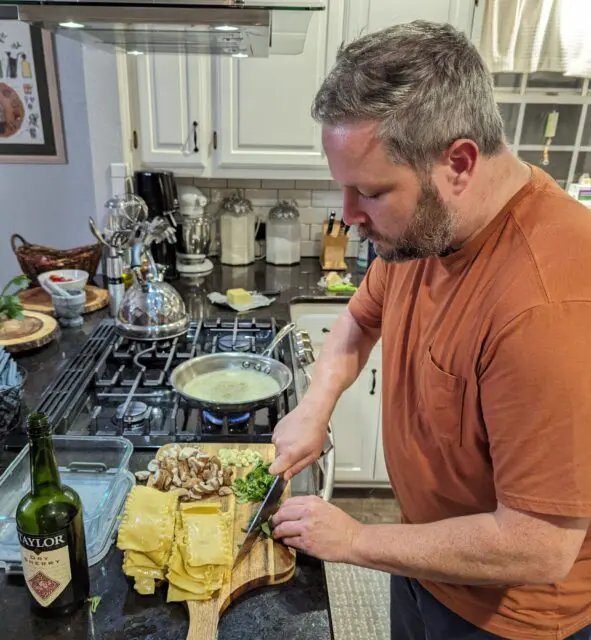 Chris chopping herbs on a cutting board with pasta, chopped walnuts, and garlic. In the background is a saute pan with lots of butter melted in it. A bottle of sherry sits nearby.