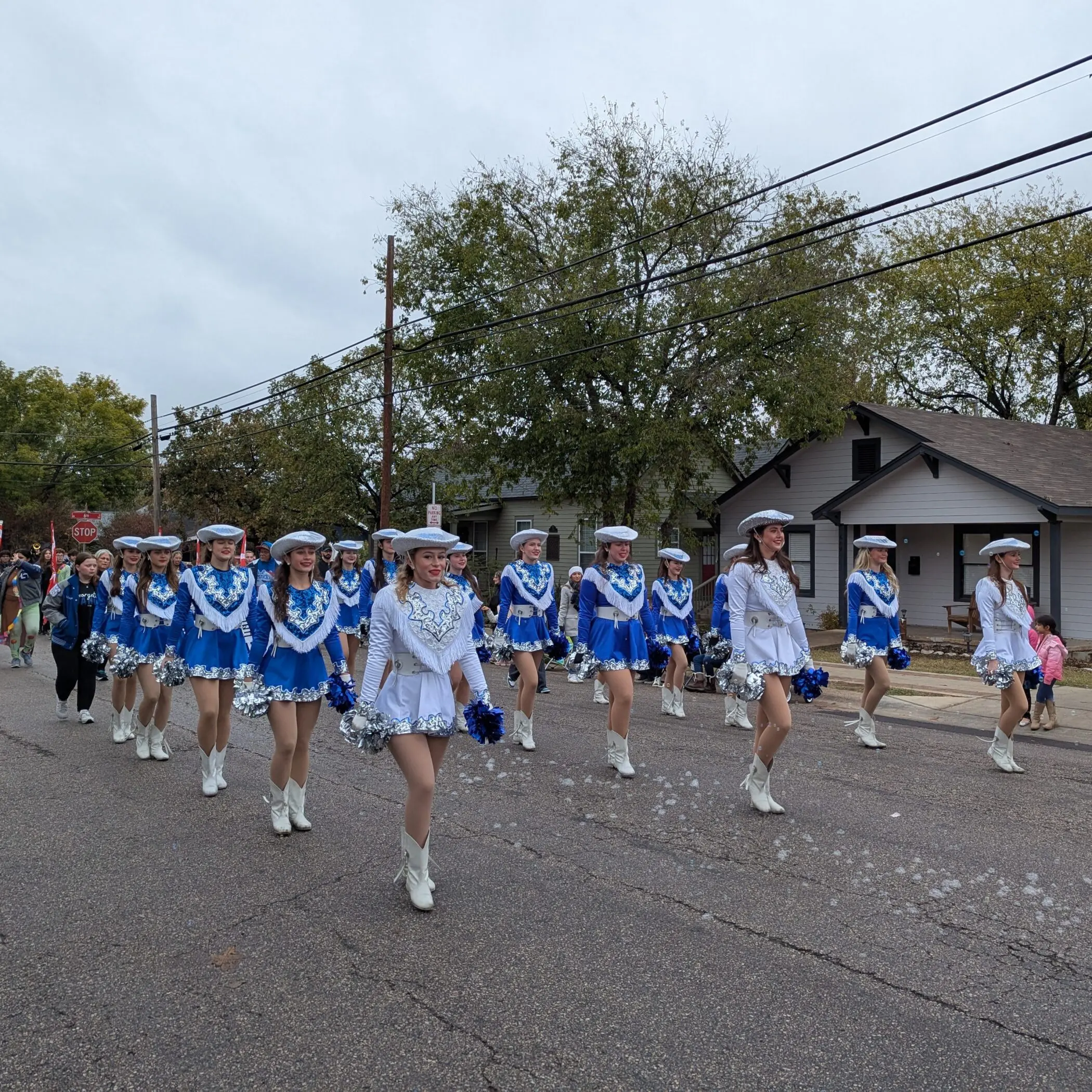 Teenage girls wearing blue, white, and silver drill team uniforms with fringe, cowboy hats and boots, and carrying silver and blue pom-poms as they march in formation.