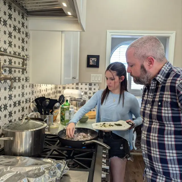 Nora putting fresh herbs in a pan with sautéed onions as Chris looks on.