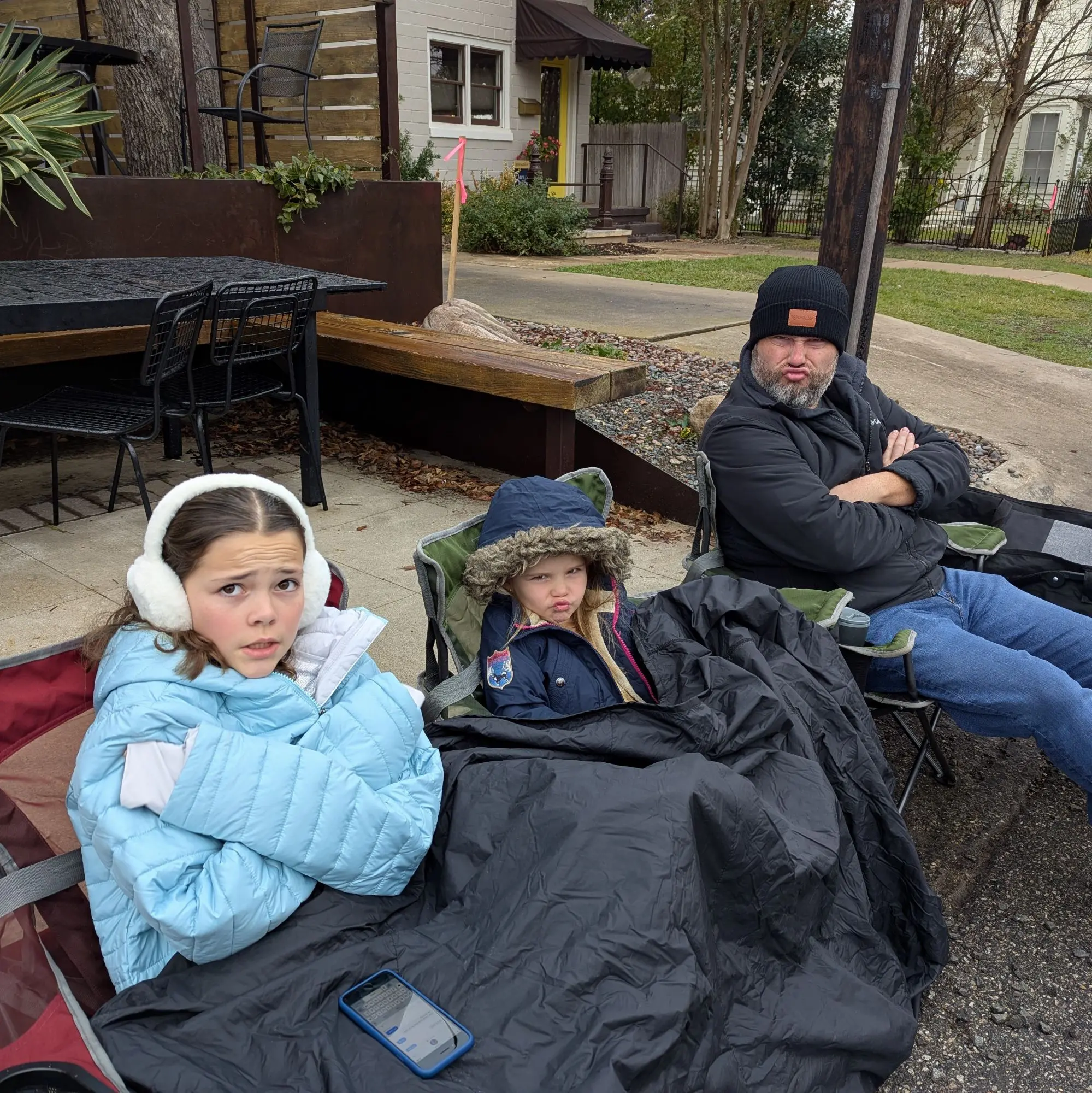 Zara, Vivi, and Chris in winter coats and hats sitting in camping chairs and making grumpy faces because it's cold.