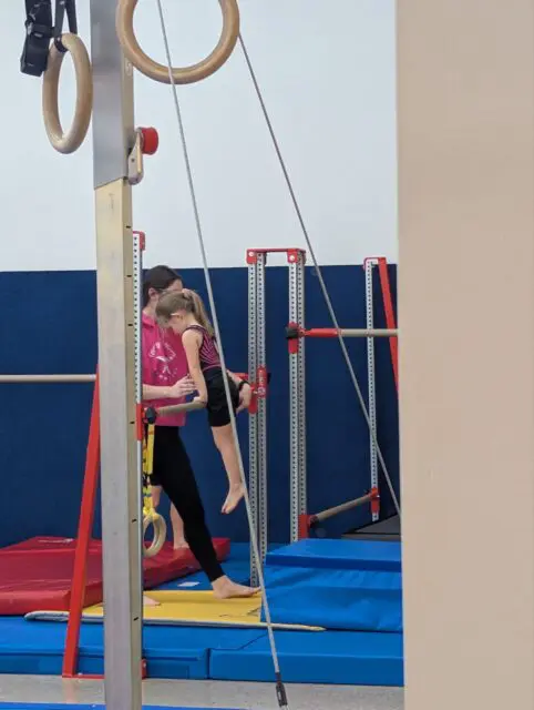 Vivi holding herself up on a bar with her arms locked and feet several feet above the floor as a teacher helps her.