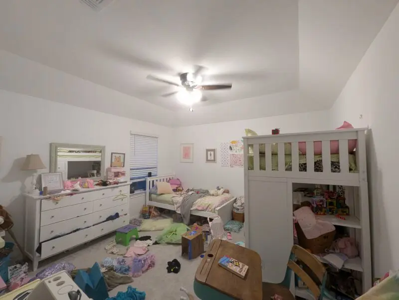 The real "before" photo showing a little girls room with two beds, a dresser, and desk, with toys and clothes covering every surface and the floor.