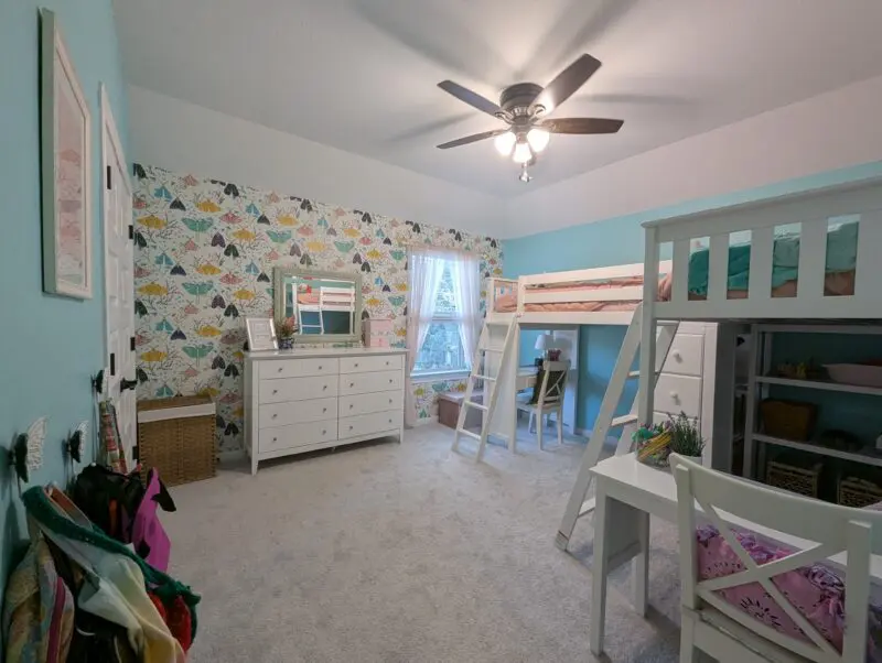 After picture, looking through the doorway into the room with bags on hooks to the left, the dresser on a wall wallpapered with yellow, pink, turquoise, and navy butterflies, and two loft beds to the right.