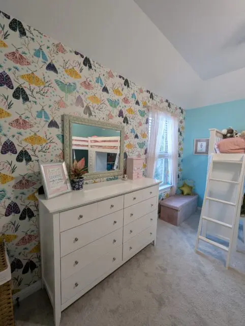 Close-up of the wallpapered wall with dresser, light turquoise framed mirror, pink curtains on the window, and a pink storage bench by the window. A P Buckley Moss print of a girl in pink with a soccer ball is hung on the turquoise wall near the window.