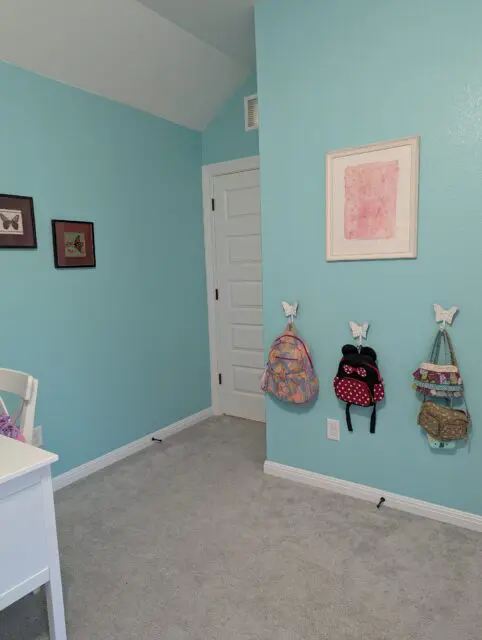 After: turquoise walls, embroidered butterfly pictures to the left of the door, butterfly hooks for bags and a pink watercolor to the right of the door.