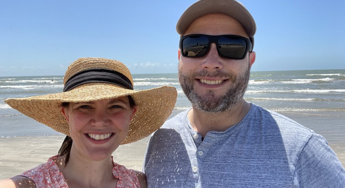 Amber wearing a sun hat and Chris with a baseball cap and sunglasses on a beach