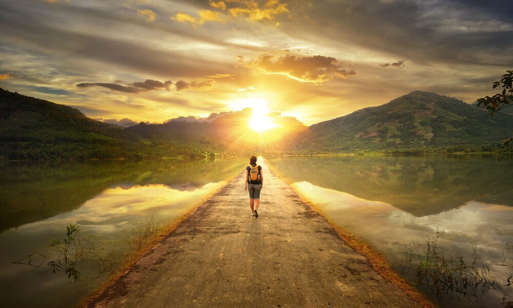 woman walking down a path surrounded by water towards a sunset over the mountains.